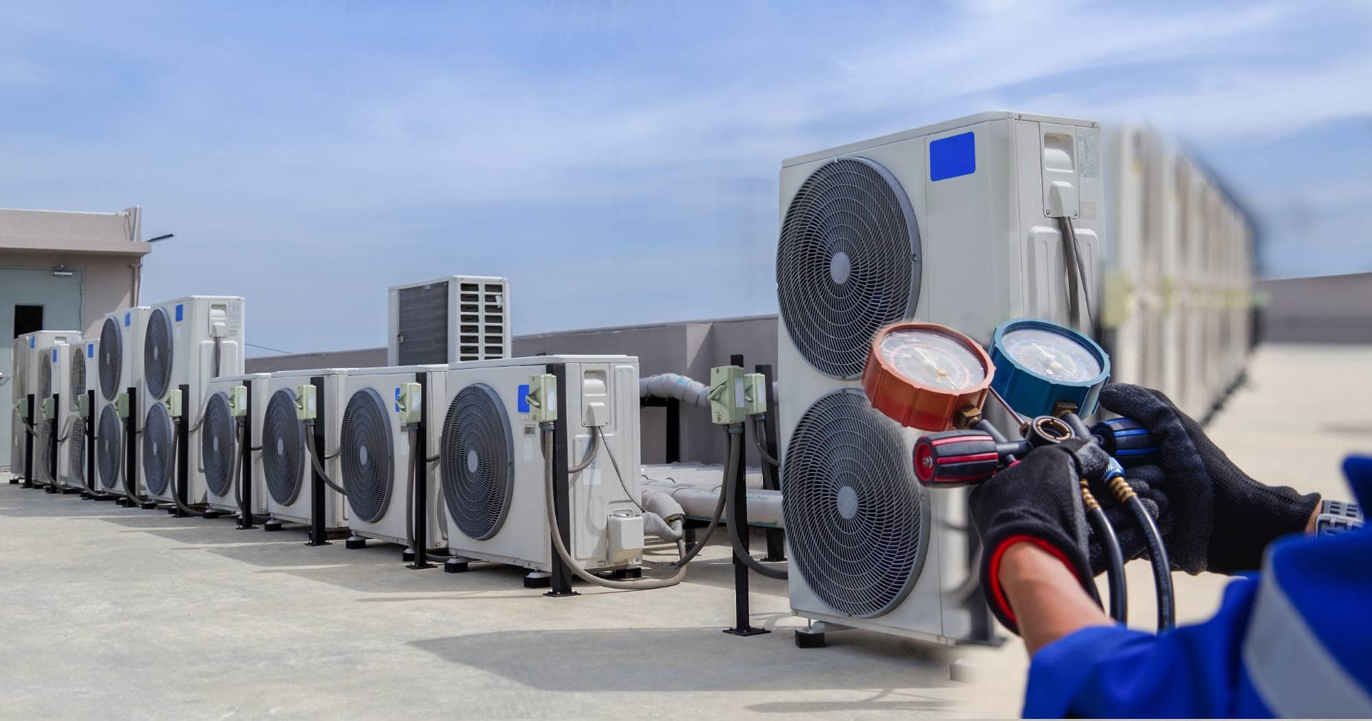A row of outdoor air conditioning units on a rooftop, with a technician&rsquo;s gloved hands holding HVAC manifold gauges in the foreground under a clear sky&mdash;serving Cooling Heating needs in San Jose & Santa Clara County, CA.