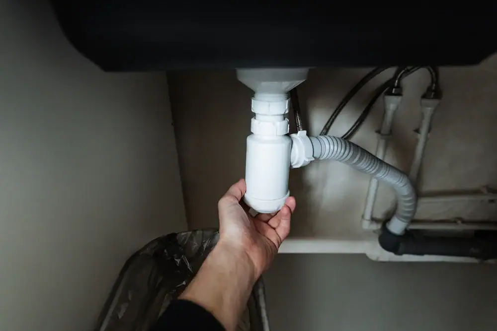 A person’s hand holds a white plastic pipe under a sink, possibly checking or repairing the plumbing beneath the basin. Various pipes and connections are visible, typical of cooling and heating systems in San Jose & Santa Clara County, CA.