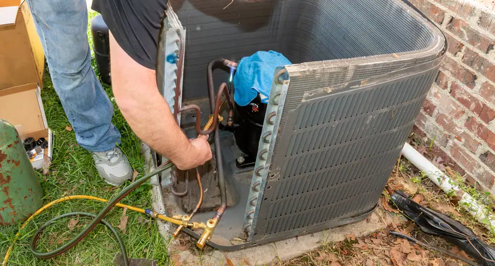 A person repairs or installs an outdoor air conditioning unit, connecting hoses to the compressor near a brick wall. Tools and equipment are visible on the ground nearby, showcasing cooling and heating work in San Jose & Santa Clara County, CA.