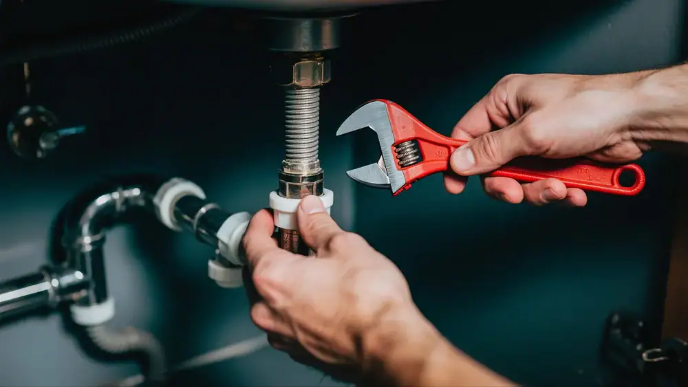 A person uses a red adjustable wrench to tighten a pipe fitting under a sink, performing plumbing work with visible pipes and connectors in CA, ideal for Cooling Heating San Jose & Santa Clara County needs.
