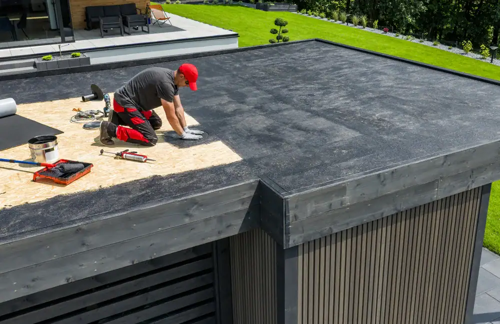 A person in a red cap and black clothing is installing roofing material on a flat roof for Cooling Heating San Jose & Santa Clara County, CA. Tools and supplies are nearby, with the building surrounded by a green lawn and landscaped garden.
