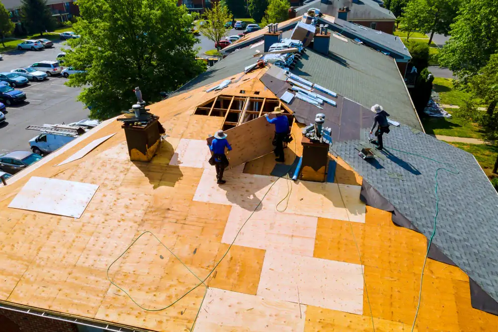 Aerial view of workers installing new plywood sheets on a large residential roof in CA, with tools and roofing materials spread out and some areas covered in black shingles. Cooling Heating San Jose & Santa Clara County services nearby homes. Trees and parked cars surround the building.