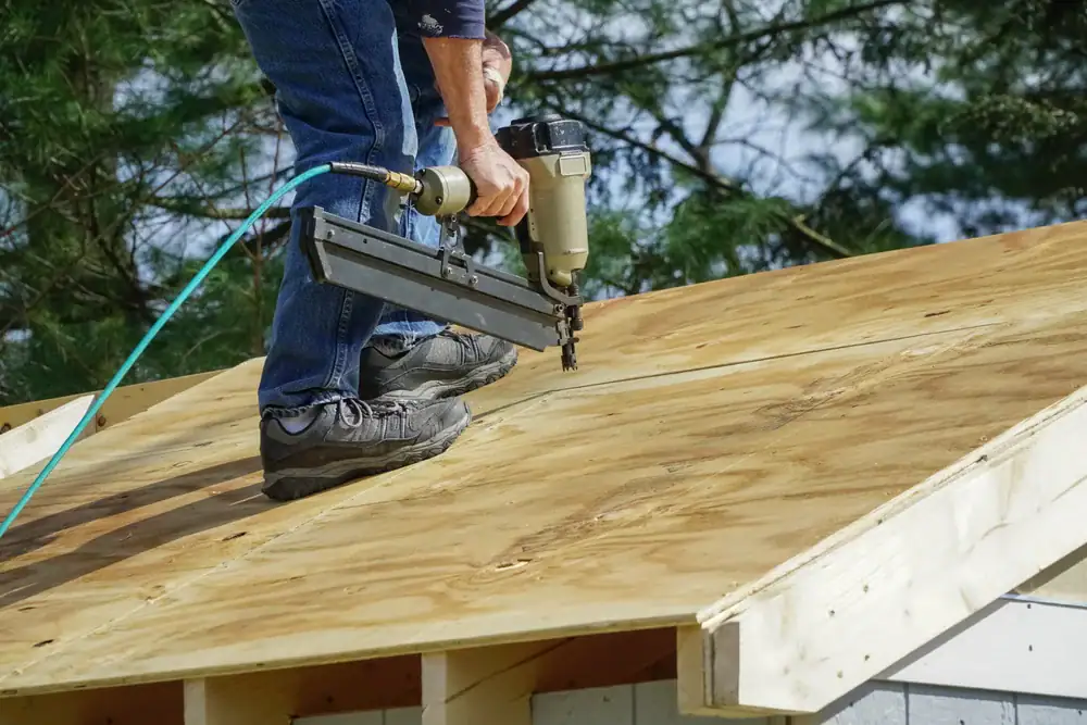 A person wearing jeans and work boots uses a nail gun to secure plywood sheets to a house roof under construction in CA, with trees visible in the background—ideal for future Cooling Heating San Jose & Santa Clara County systems.