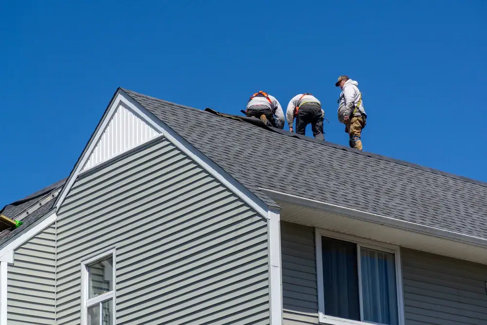 Three workers in safety gear are installing or repairing shingles on the sloped roof of a gray house with white trim, under a clear blue sky—typical scenes for Cooling Heating San Jose & Santa Clara County, CA home improvements.