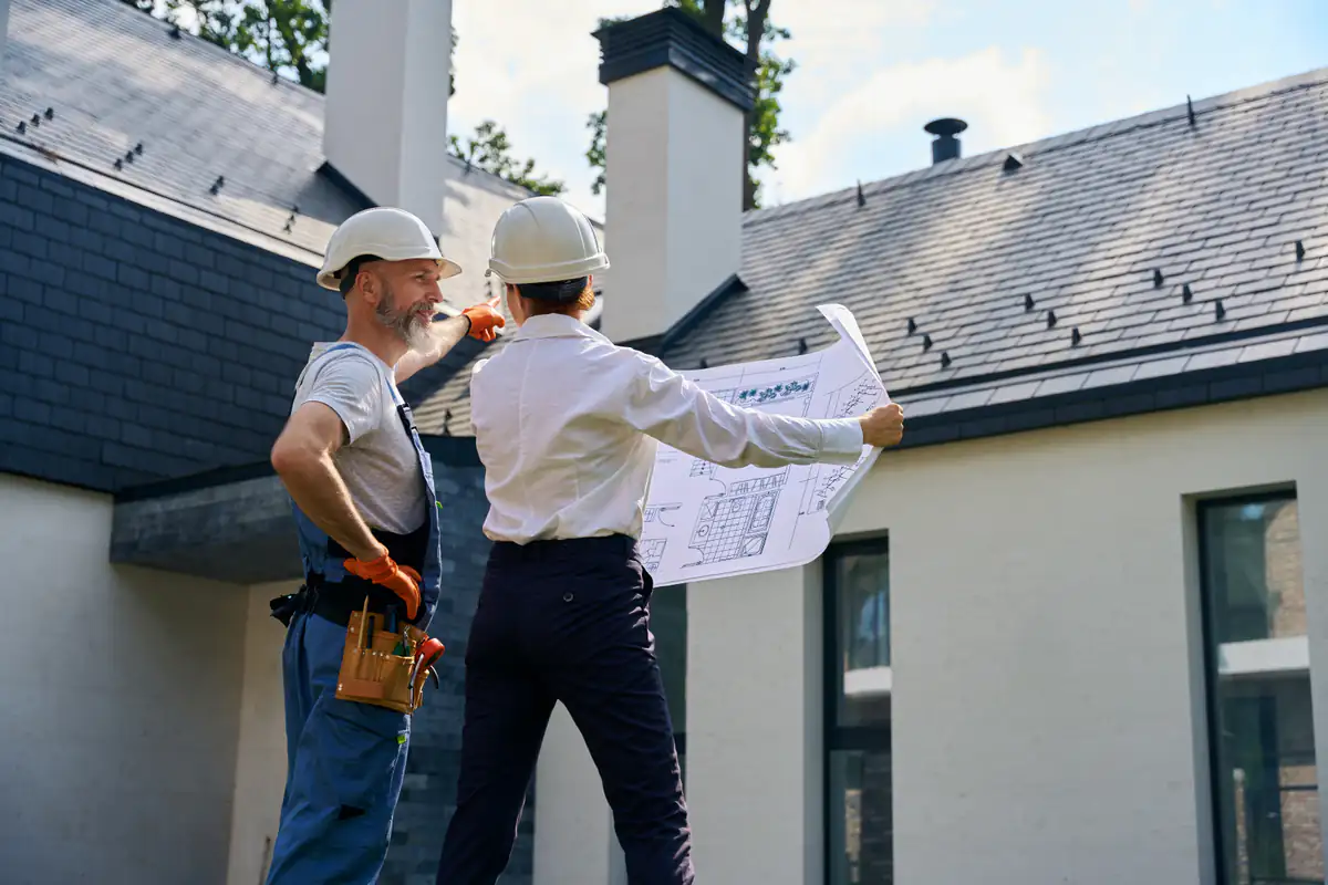 Two construction workers in hard hats stand outside a modern house; one holds blueprints while the other points toward the roof, appearing to discuss plans or repairs.