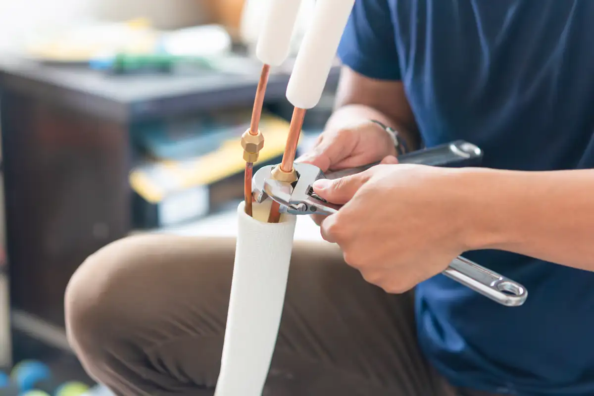 A person uses a wrench and pliers to tighten copper pipes covered with insulation, likely working on an air conditioning or refrigeration unit.