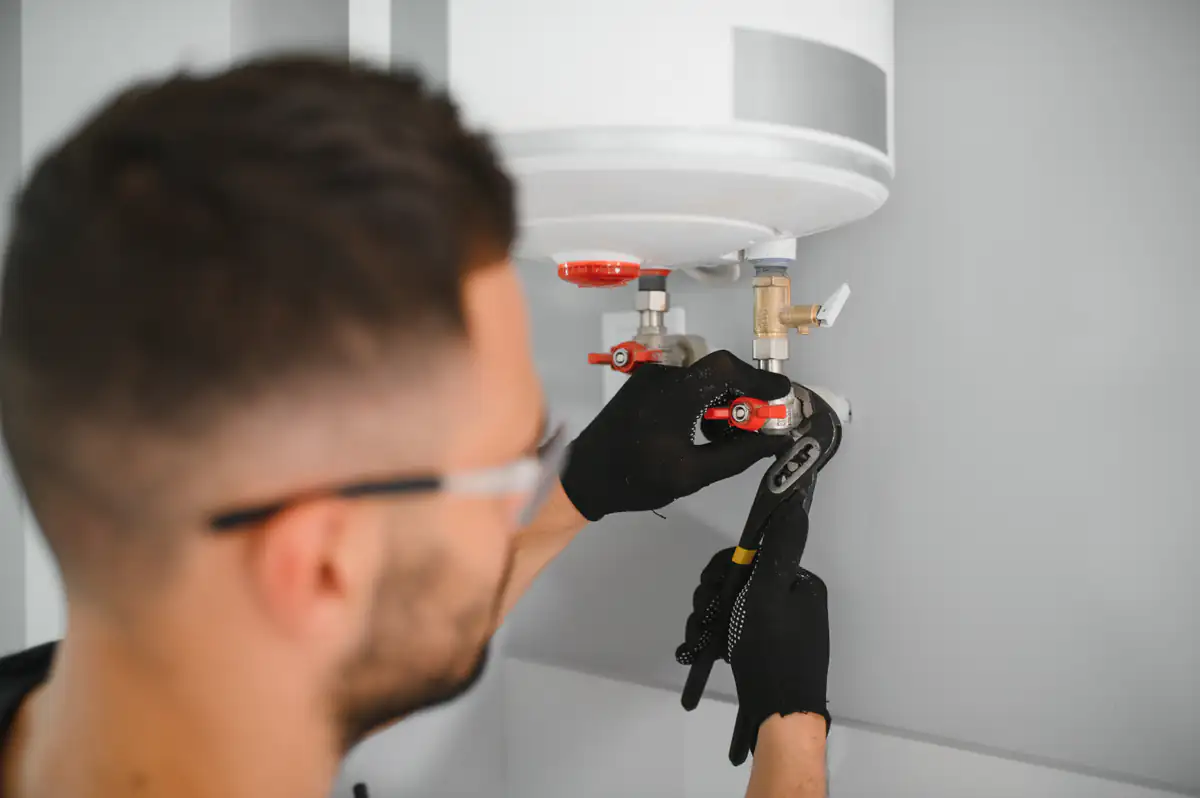 A person wearing safety glasses and black gloves uses a wrench to adjust valves under a white water heater mounted on a wall.