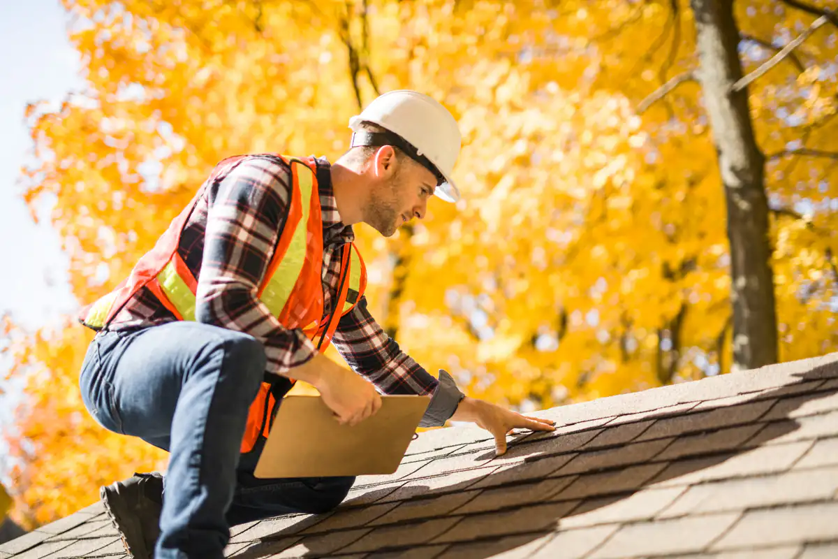 A construction worker wearing a hard hat, safety vest, and plaid shirt inspects roof shingles on a house, with bright yellow autumn trees in the background.