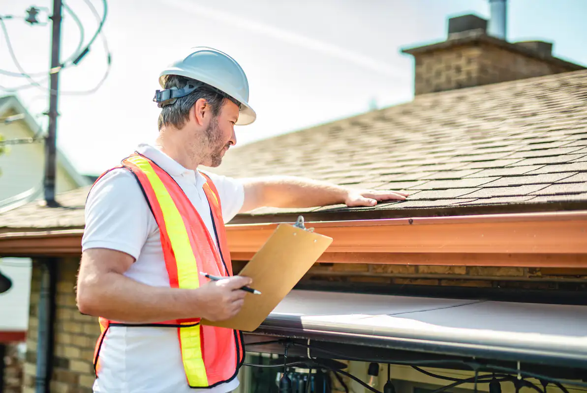 A construction worker wearing a hard hat and reflective vest inspects roof shingles on a house while holding a clipboard and pen.