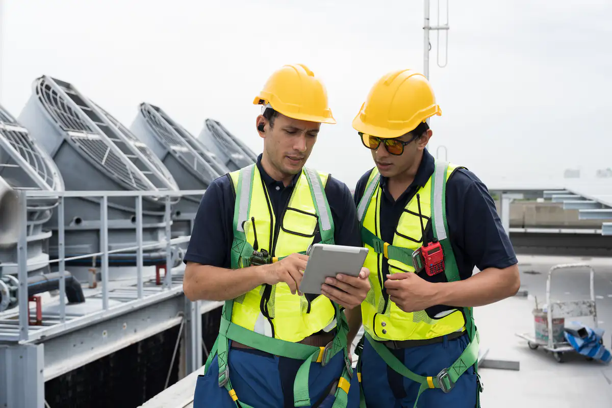 Two construction workers wearing yellow hard hats and safety vests stand on a rooftop, looking at a tablet. Large industrial fans are visible in the background.