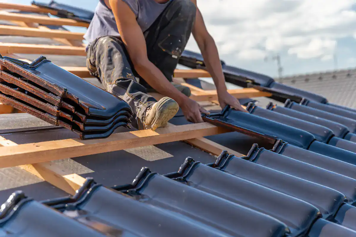 A worker installs dark roof tiles on a wooden frame atop a building under construction, with stacks of tiles and blue sky in the background.
