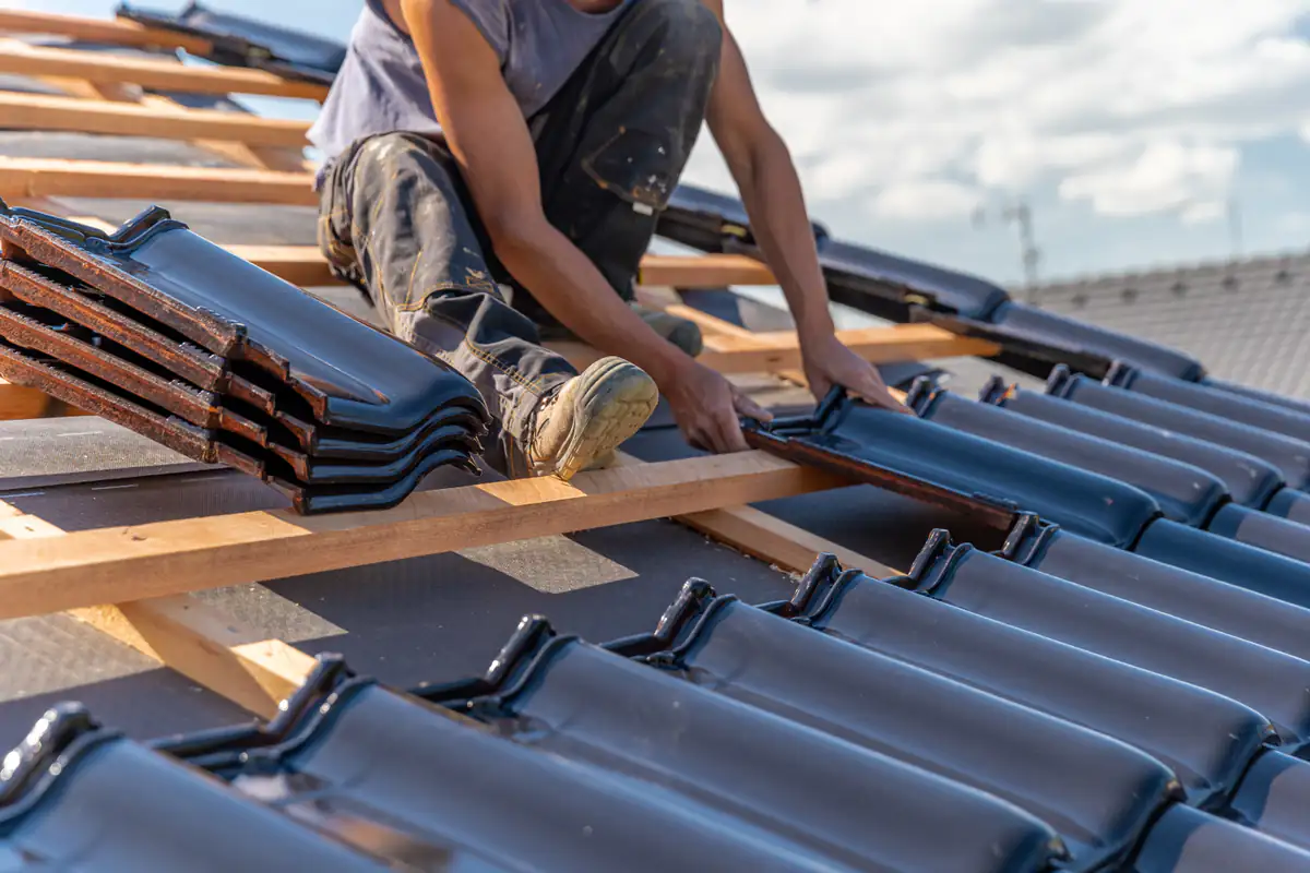A person installs dark roof tiles on a wooden frame of a house roof under a partly cloudy sky, with stacks of tiles nearby.