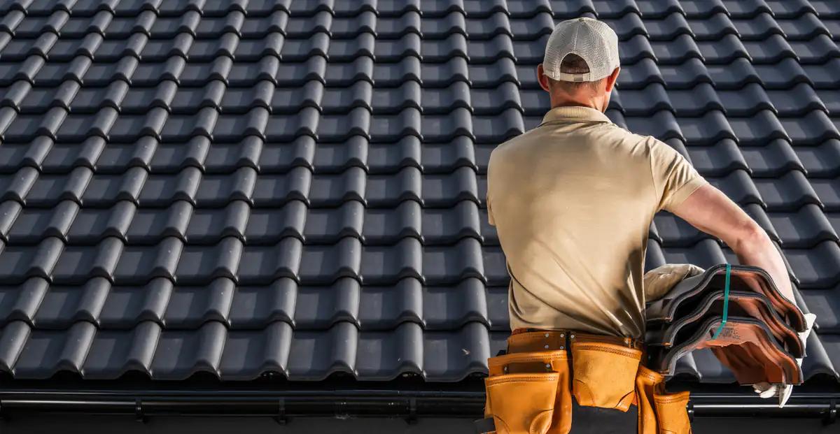 A worker wearing a cap and tool belt stands on a roof, holding a bundle of roof tiles, facing a large expanse of gray, neatly arranged roof tiles.