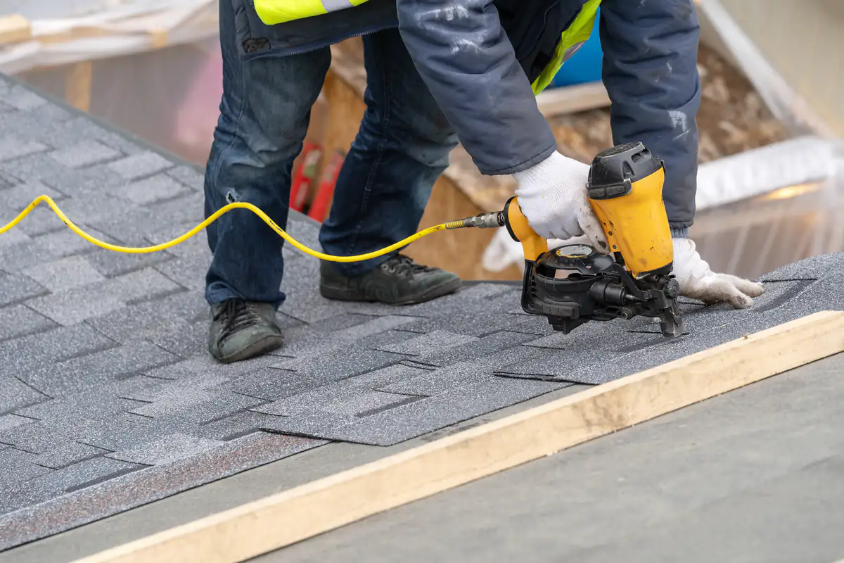 A worker in protective gloves and a safety vest uses a yellow nail gun to install asphalt shingles on a sloped roof, with wooden boards and construction materials visible nearby.