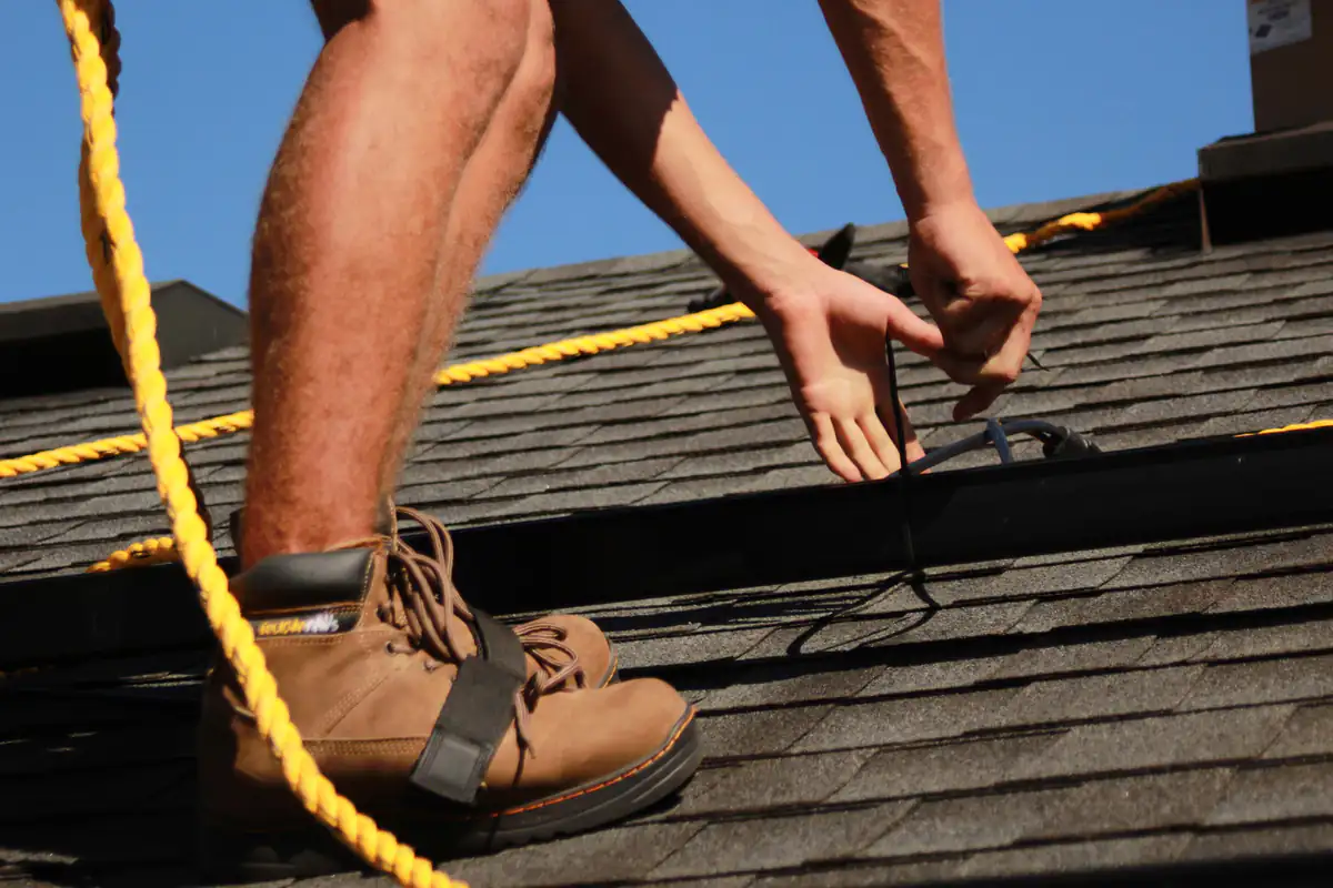 Close-up of a person wearing brown work boots installing equipment on a shingled roof, with yellow safety ropes visible and hands securing black wires against a clear blue sky.