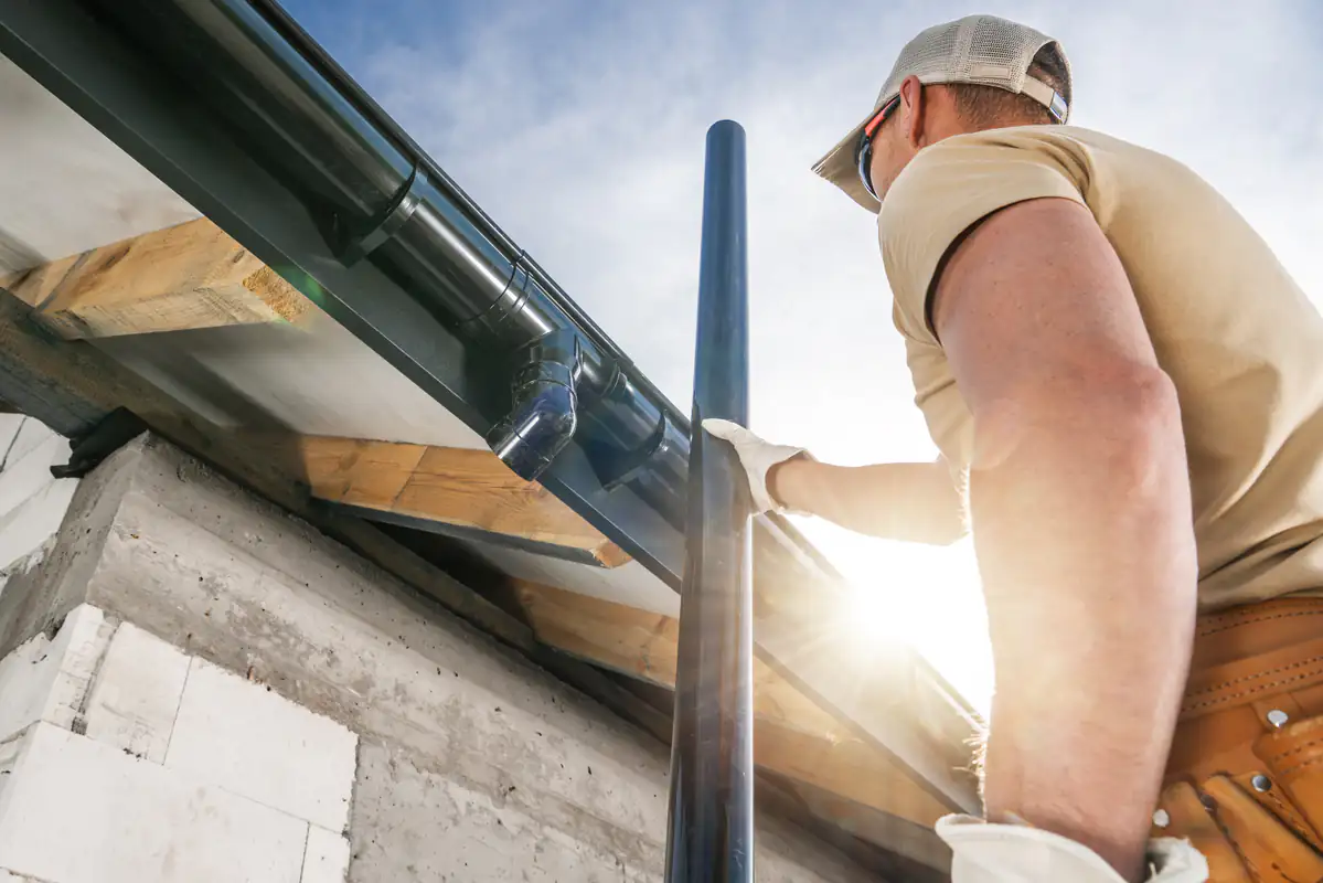 A construction worker wearing gloves and a cap installs a black downspout onto a roof gutter of a building under construction, with the sun shining in the background.