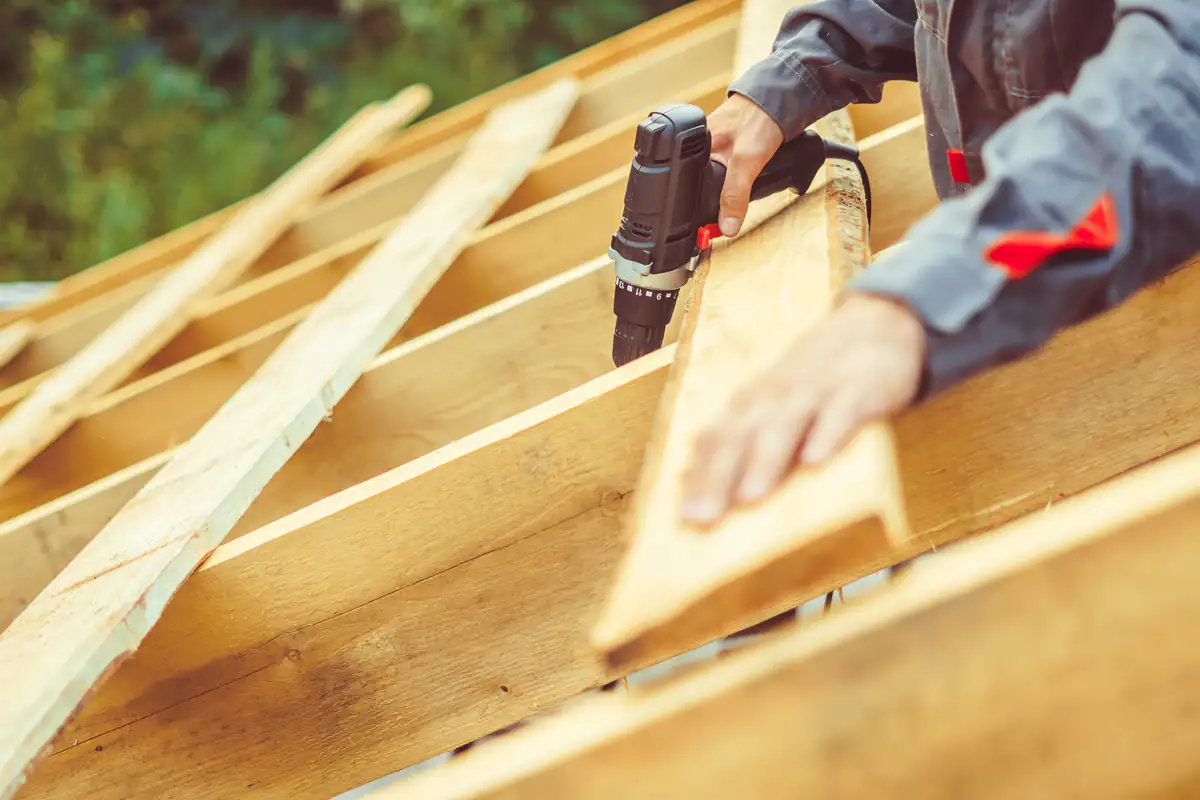 A person wearing a gray work jacket uses a cordless drill to screw a wooden plank onto a roof frame under construction. The background is blurred with greenery.